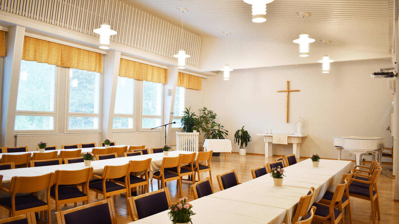 Kurun seurakuntatalon sali, jossa on pitkiä pöytiä ja niiden ääressä tuoleja sekä päätyseinällä risti.