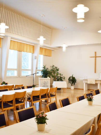 Kurun seurakuntatalon sali, jossa on pitkiä pöytiä ja niiden ääressä tuoleja sekä päätyseinällä risti.