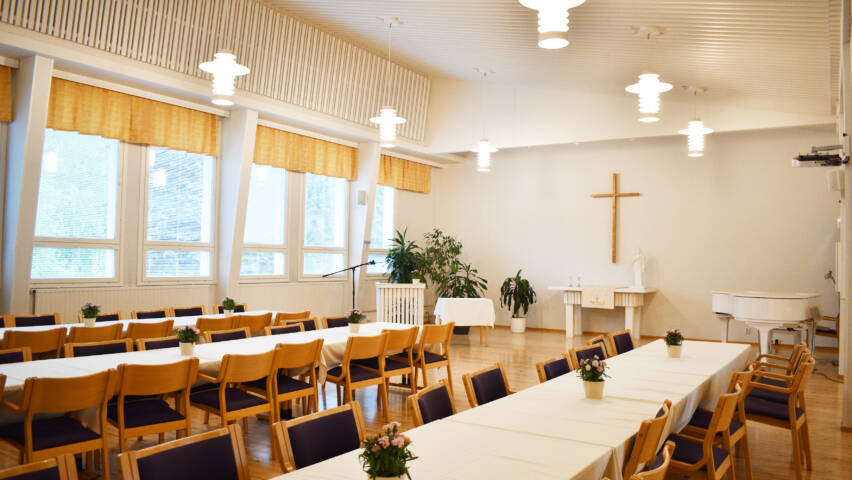 Kurun seurakuntatalon sali, jossa on pitkiä pöytiä ja niiden ääressä tuoleja sekä päätyseinällä risti.