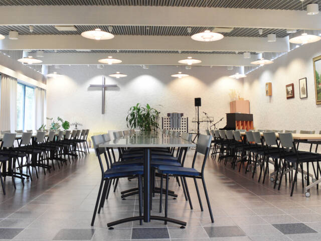 Viljakkalan seurakuntatalon sali. Pitkien pöytien toisessa päässä seinää koristaa iso risti, jonka edustalla on puhujankoroke ja mikrofonitelineitä.