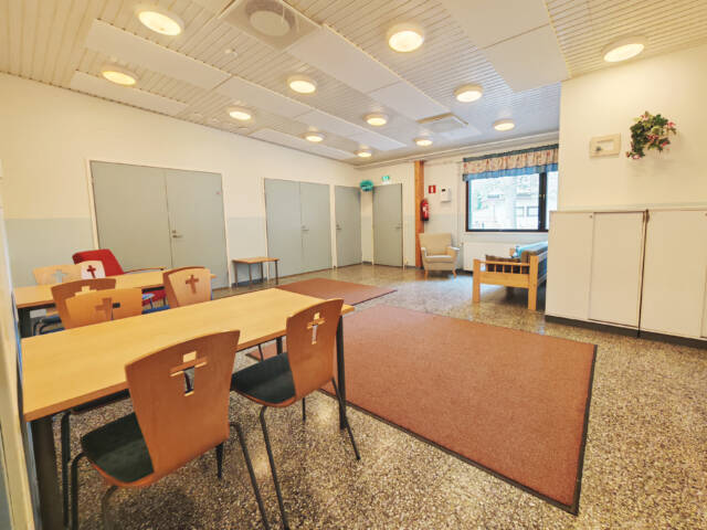 Vuorentaustan seurakuntatalon huone. Lattialla on matto, seinustalla pöytiä ja tuoleja sekä ikkunan edessä istumapaikkoja.