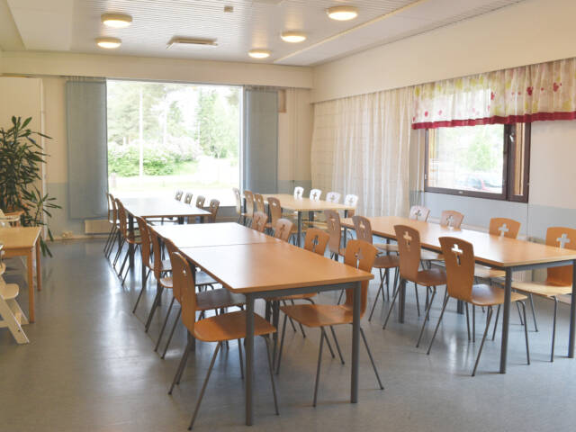 Vuorentaustan seurakuntatalon ruokailutila, jossa on ikkunan edustalla pitkiä pöytiä ja niiden ääressä tuoleja.