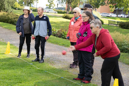 Sivusta kuvattuna kaikkiaan kuusi ihmistä seisomassa rintamasuunta samaan suuntaan. Yksi heittää juuri nurmikolle boccia-palloa.